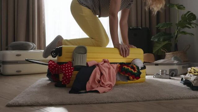 Woman trying to close suitcase sitting on it. Woman packing suitcase before travel trip flight. Too many lot of clothes in suitcase baggage not closing not zipping luggage. Clothes packing before trip