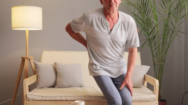 Unhealthy gray haired man suffering from strong pain in back after abruptly rose from the sofa touching painful place frowning face from injury.