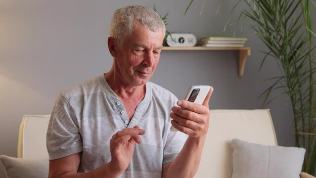 Happy Caucasian man wearing casual T-shirt using smartphone device while sitting on sofa at home reading messages on mobile phone scrolling online browsing web pages.