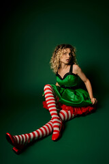 dreamy curly woman dressed as new year elf sitting on floor posing on dark green background