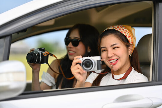 Two excited female tourists taking photos from a car window with vintage retro cameras during their summer road trip