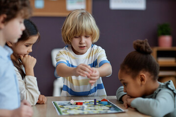 Diverse group of little kids playing board game together in preschool and throwing dice, copy space