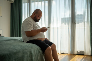 Overweight guy hanging in smartphone on resort sitting in bed in hotel room on vacation. Man has...