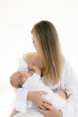 Obraz na płótnie Canvas Young woman with baby in her arms. Blonde girl in white clothes smiling to your child on white background. Happy motherhood and breastfeeding concept. Photos with sun glare, soft focus, overexposure