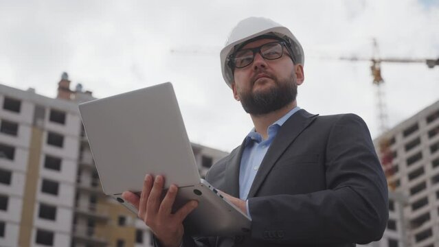 Bearded civil engineer in suit and protective helmet making important notes in notebook about the work done at construction site. Low angle man inspecting construction site. Concept of control