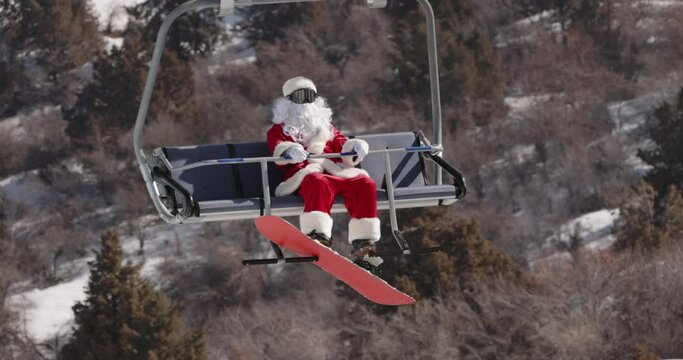 Santa Claus snowboarder on winter cable car in mountains. Ski holidays. Snowboard lift and gondola. Winter sport and outdoor activities. Man in Santa suit rides up mountain on funicular to snowboard