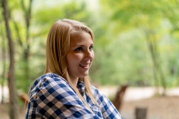 A young smiling blonde girl in a blanket warms herself by the fire near her wooden house in forest