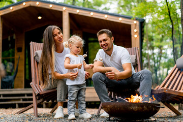 Cute young family dad mom and daughter are sitting together by fire in the forest and roasting marshmallows near their country house