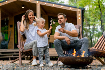 Cute young family dad mom and daughter are sitting together by fire in the forest and roasting marshmallows near their country house - Powered by Adobe