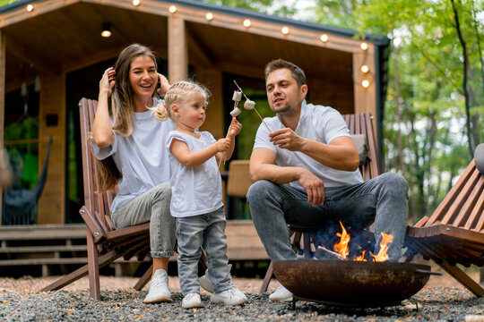 Cute young family dad mom and daughter are sitting together by fire in the forest and roasting marshmallows near their country house