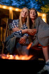 Girlfriends in blankets sit in the evening by the fire near country wooden house in the forest