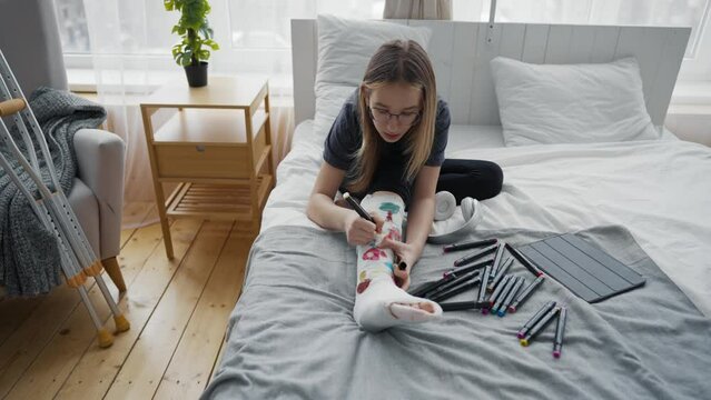 Teen girl painting colorful pictures using felt-tip pens on orthopedic cast on broken leg sitting in bed at home. Boring teenager resting relaxing drawing. Recovery of damaged leg, sick leave concept.