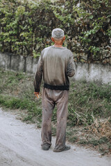 An old adult gray-haired thin homeless hungry man in dirty clothes walks along the street road in...
