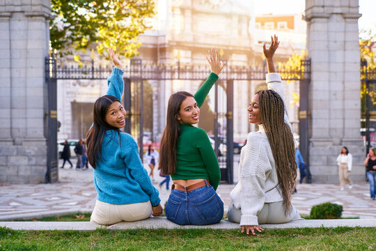 Group Of Three Multiracial Women Friends Sitting Outdoors In The City Turning Back To Look At Camera Cheerfully And Raising Hands, Gathering Together After University.