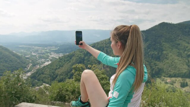 Hiking girl sitting on a bench at a viewpoint enjoying taking photos of beautiful mountains and valley landscape with a smartphone, rear view.