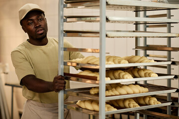 Portrait of Black young man moving shelf with baked goods in bakery kitchen, copy space