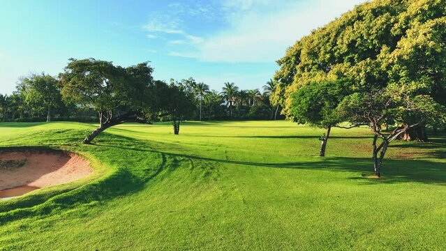 Beautiful evening landscape of a golf club with rich green turf. Panoramic view of the golf course with green grass lawn and tropical trees. Sand trap in a meadow.