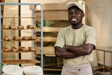 Waist up portrait of Black young man standing in bakery kitchen with arms crossed and smiling at...