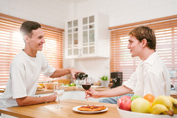 Caucasian gay attractive couple pouring and drinking red wine together in the home kitchen with fun and smiling. Homosexuality, same-sex relationships, LGBT, gay partner and romance concept.