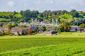 Esplanade with green grass on Caestert plateau, suspension bridge, Kanne village and hill with green leafy trees in background, Belgian agricultural land with sown land, sunny day in Riemst, Belgium