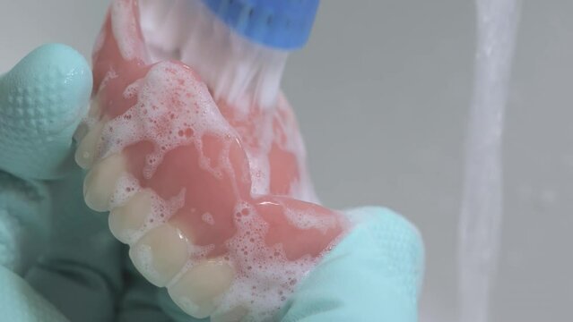 Closeup shot of cleaning prosthesis teeth using a brush. Rubber gloves being used. Tap water in the background