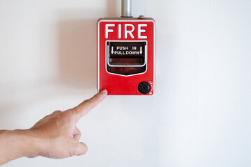 Emergency fire warning system pull station. Hand pointing at red fire alarm mounted on white wall in public building