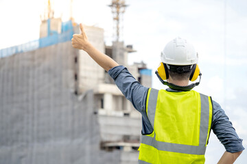 Construction worker man with green reflective vest and safety helmet showing thumbs up at unfinished building structure and tower crane. Male site engineer showing hand gesture sign for approval - obrazy, fototapety, plakaty