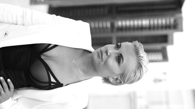 Fashionable blonde with short hair and sunglasses in a white jacket on the street on a sunny day. Black and white vertical video.