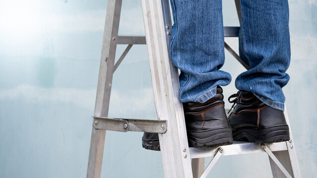 Male maintenance worker feet with protective safety shoes climbing up on aluminum step ladder at construction site. Building service tool and equipment