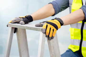 Male maintenance worker hands with protective yellow gloves holding aluminum step ladder at construction site. Building service tool and equipment - obrazy, fototapety, plakaty