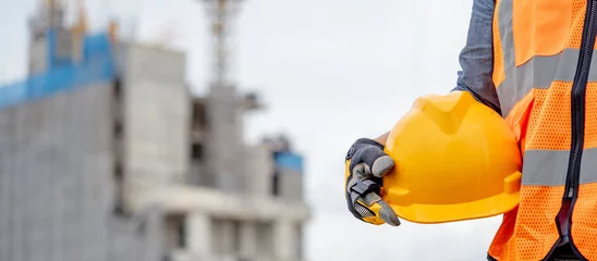 Fotobehang Safety workwear concept. Male hand holding yellow safety helmet or hard hat. Construction worker man with reflective orange vest and protective gloves standing at unfinished building with tower crane © Summer Paradive