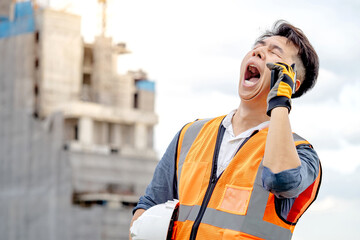 Asian male construction worker with orange reflective vest yawning while using phone at construction site. Male engineer feeling sleepy and tired from insomnia or insufficient rest - obrazy, fototapety, plakaty