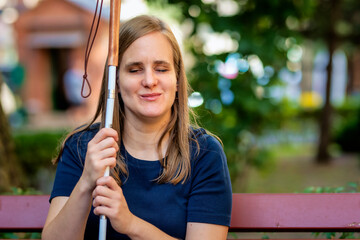 A visually impaired woman holding a white cane and sitting on the bench on the city park