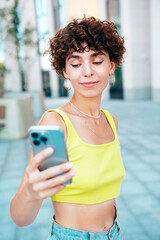 Young beautiful smiling hipster woman in trendy summer clothes. Carefree woman with curls hairstyle, in the street. Positive model holds mobile phone, looks at cellphone screen, uses smartphone apps © halayalex
