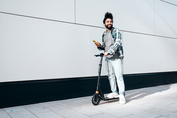 Trendy smiling bearded man in casual clothes riding electric scooter in urban background. Handsome model posing in the street near wall. Hipster guy using sharing system application on his smartphone