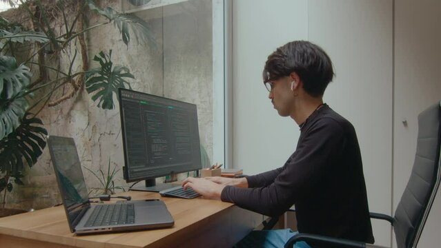 Male computer programmer in wireless earphones coding both on desktop PC and laptop in modern home office. Zoom shot