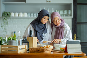 muslim mother looking at recipe from laptop and cooking with her daughter. having fun woman with hijab and kid preparing dinner together, Islamic Lady With Small Daughter Enjoying Doing Homemade