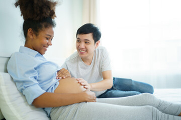 Happy cheerful husband and pregnant wife staying - relaxing together in bedroom, husband embracing or playing on pregnant wife tummy. Diverse ethnicity husband and wife lifestyles concept.