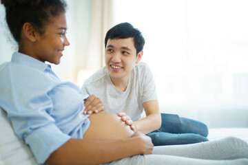 Happy cheerful husband and pregnant wife staying - relaxing together in bedroom, husband embracing or playing on pregnant wife tummy. Diverse ethnicity husband and wife lifestyles concept.