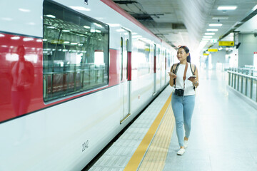 Happy beautiful Asian woman waiting for a train at railway platform. Lonely Asian female tourist or traveler standing at train station. - obrazy, fototapety, plakaty