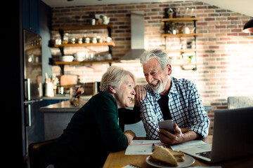 Senior couple using a smartphone while going over financials in the kitchen - obrazy, fototapety, plakaty