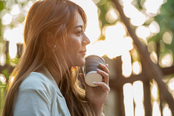 Asian woman holding hot coffee in paper mug cup to sniff smell of espresso in morning sunlight....