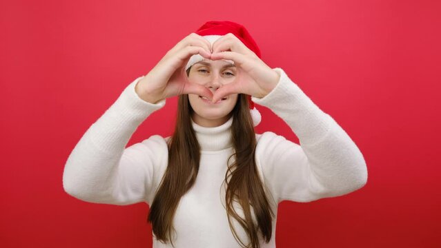 Merry cute young woman in cozy sweater and Xmas Santa hat showing shape heart look through hands heart-shape sign, posing isolated over pastel red background wall. Happy New Year 2024 holiday concept