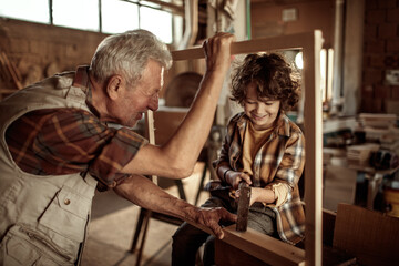 Fototapeta na wymiar Senior male carpenter teaching his grandson how to use a hammer in a carpentry shop