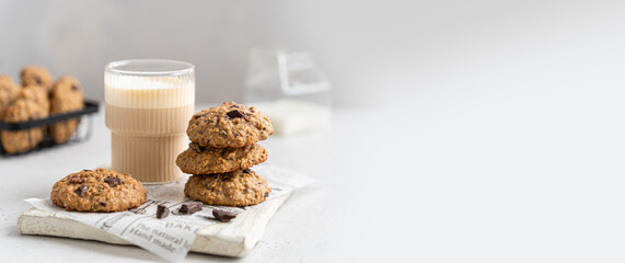 Homemade oatmeal cookies banner. Oatmeal biscuits with banana, oats, chocolate drops and nuts on a wooden board and a glass of latte on a white background. Copy space. Bakery web line. Breakfast