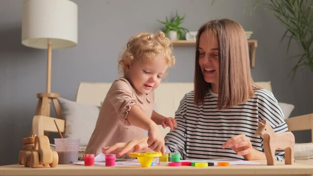 Smiling cheerful mommy with her small daughter painting together in light room interior drawing with pain and fingers enjoying funny and creative process of having fun and education.