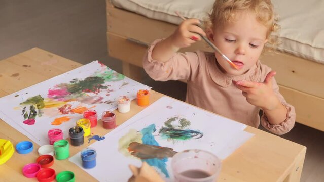 Funny blonde baby girl learning painting with water colors in light room interior on her fingers making manicure with diligent facial expression enjoying creative time.