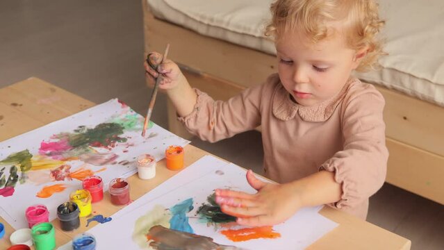 Infant painting fun. Cute funny toddler child drawing using colorful paints and brush at home or nursery making picture with painted palms making creative picture.