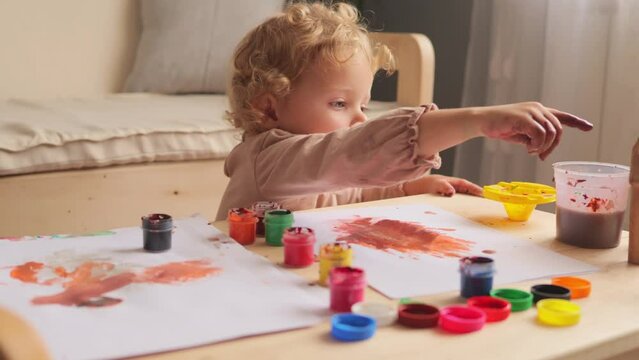 Cute diligent baby girl learning painting with water colors and fingers developing fine motor skills in light room interior little painter's artwork beautiful display of child creativity..