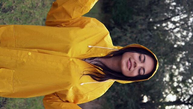 portrait of a smiling young woman in a yellow raincoat during the rain in the park. Cheerful woman enjoying the rain outdoors and dancing. vertical video